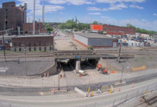 Image shows rail bridge construction, railroad tracks, buildings, sky.
